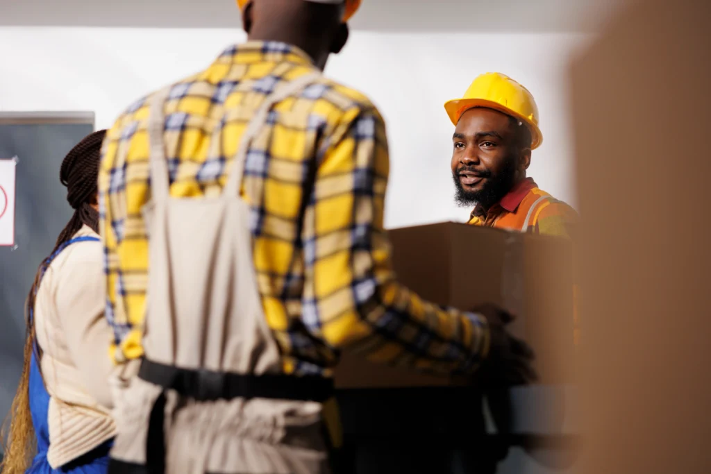 Warehouse Manager Looking Colleague Bringing Parcel Reception Desk Storage Room African American Storehouse Employee Wearing Protective Hard Hat Receiving Box From Package Handler 1024x683